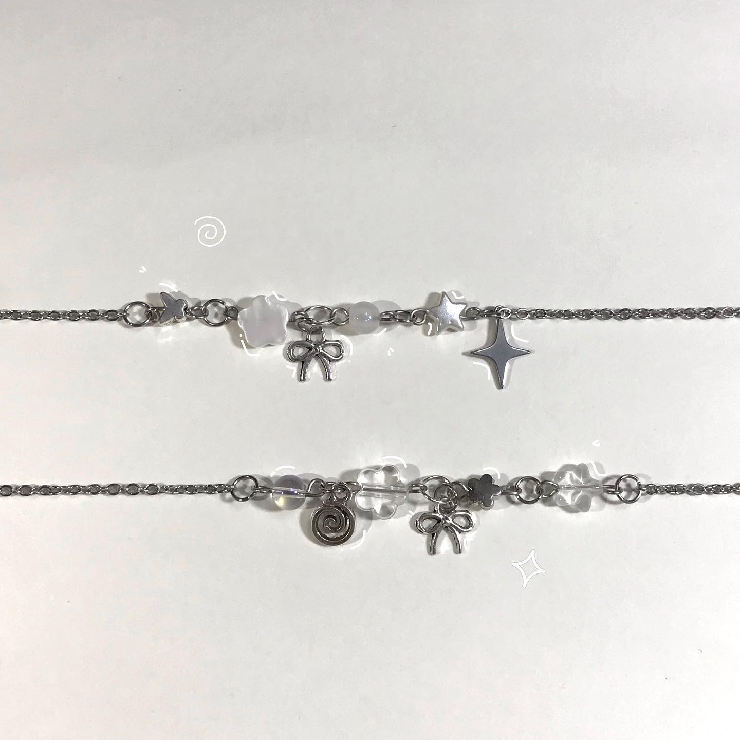 two silver bracelets with beads and various charms are displayed on a white surface. the top bracelet has a star charm and a bow charm, while the bottom one features a spiral charm and another bow charm. both bracelets are interspersed with beads.