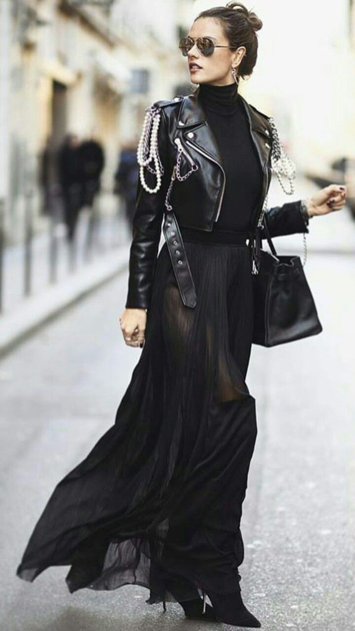 a woman wearing a black leather jacket, a long black skirt, sunglasses, boots, and a bag walks down a street in paris. she has pearls around her jacket.  she is looking towards the right side of the frame.  her skirt is sheer and has pleats.