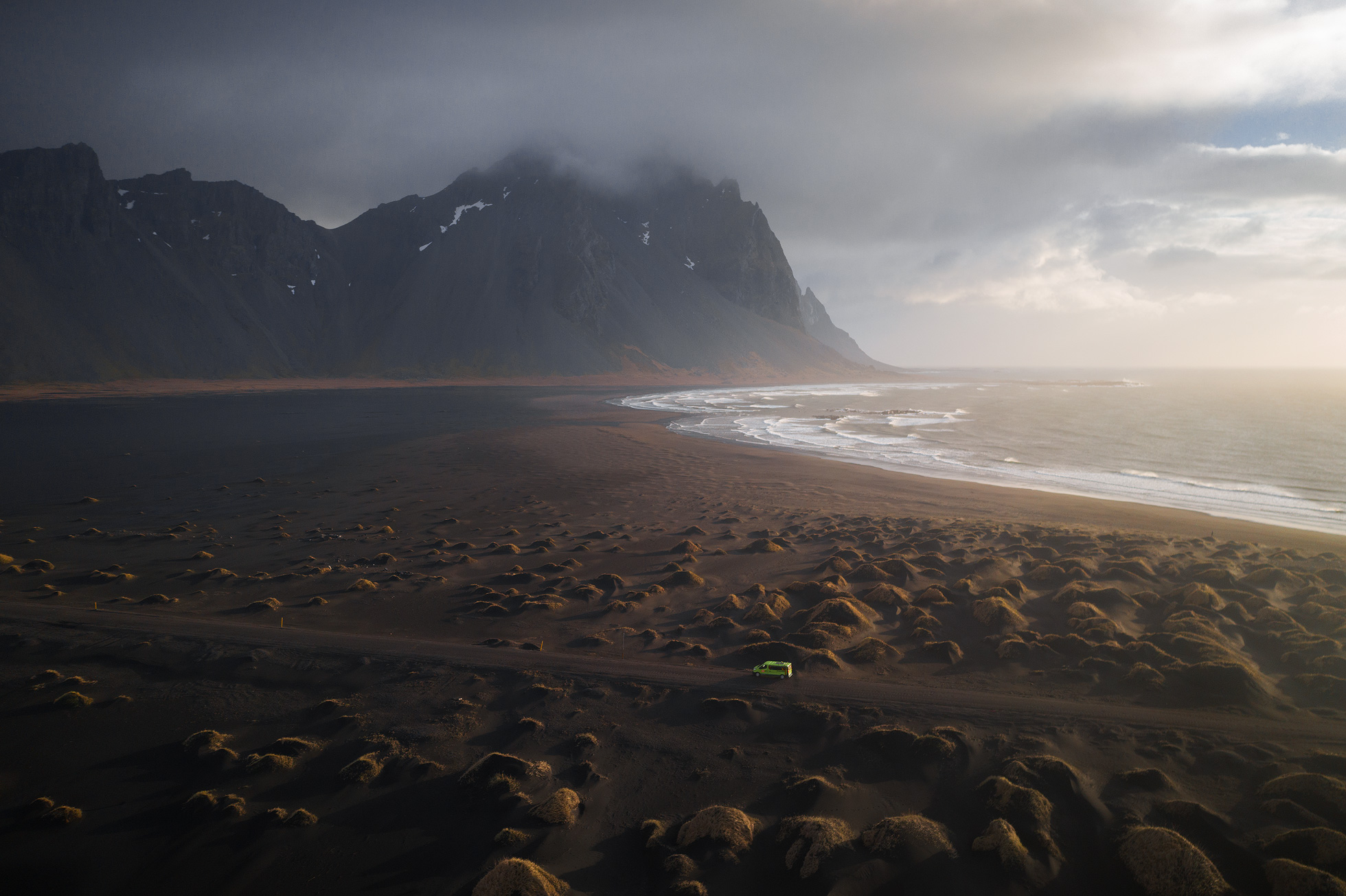 an aerial view of a desolate beach with a lone vehicle on a road. the beach is sand and dark in color. there are mountains in the background and dark, stormy looking clouds above. the ocean is calm and the sky is blue with patches of clouds. this image is a beautiful example of how powerful and dramatic nature can be.