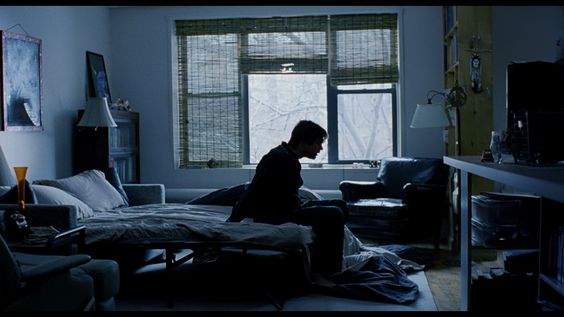 a man sits on the edge of a bed in a dimly lit apartment. the room is sparsely furnished, with a sofa, a chair, and a table in the background. a large window reveals a glimpse of bare trees outside. the man's posture suggests a sense of dejection or contemplation.  the overall atmosphere is one of solitude and introspection.
