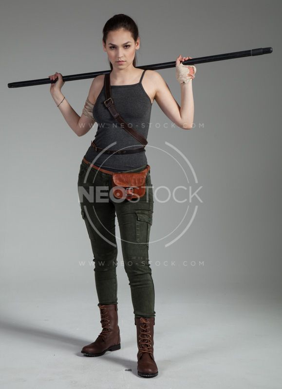 a young woman poses for a studio portrait, wearing a grey tank top and green pants, holding a metal pole on her shoulder, looking directly at the camera with a serious expression. she is wearing a belt with a bag, and brown boots. the background is grey.