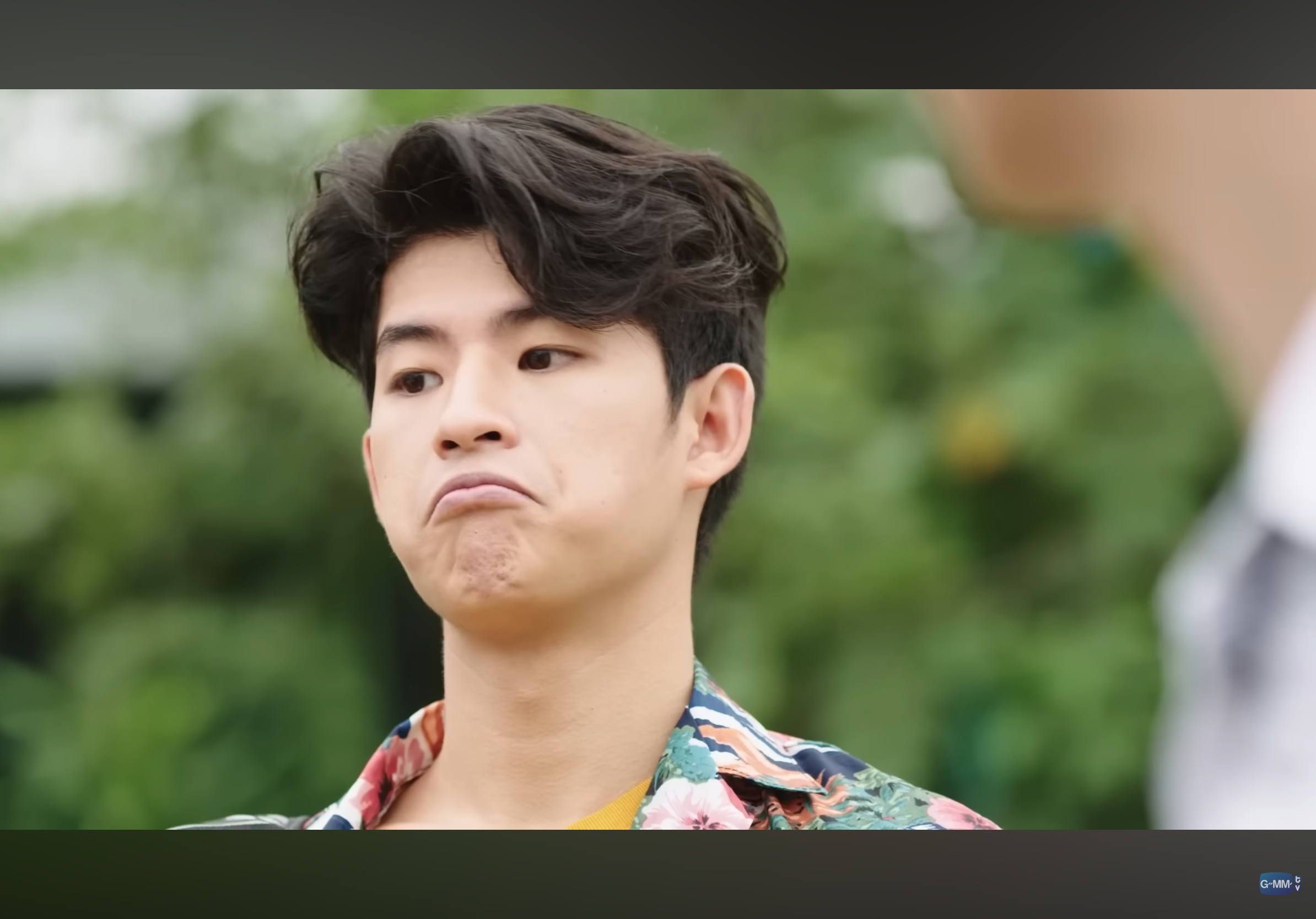 a closeup portrait of a young, asian man with a sad or disappointed expression on his face. the man has short, dark hair and is wearing a floral shirt. the background of the image is a green, outoffocus natural setting, suggesting the photo was taken outdoors. the man appears to be pouting slightly, with his bottom lip protruding.