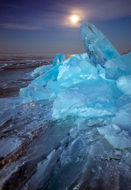 the image shows a landscape of ice chunks under the night sky with the moon shining above. the ice has a blueish tint, suggesting extreme cold and the landscape is frozen.