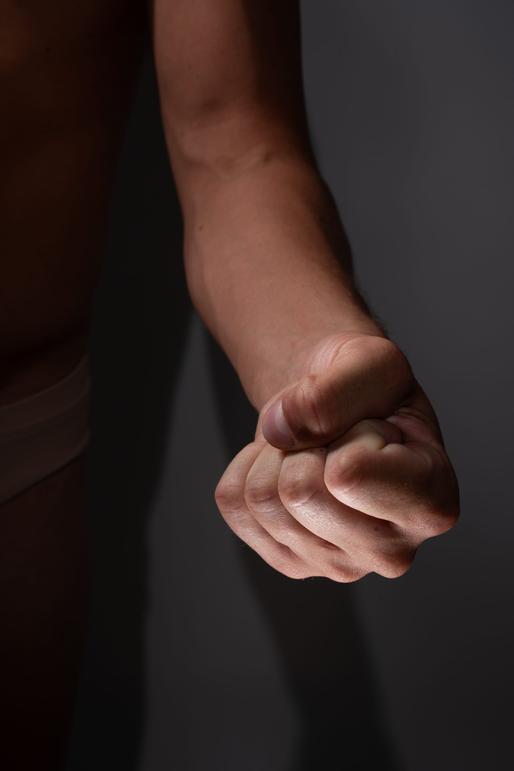 a closeup shot of a man's clenched fist, illuminated by indoor lighting against a dark gray background. the image emphasizes the tension and strength conveyed by the clenched fist.