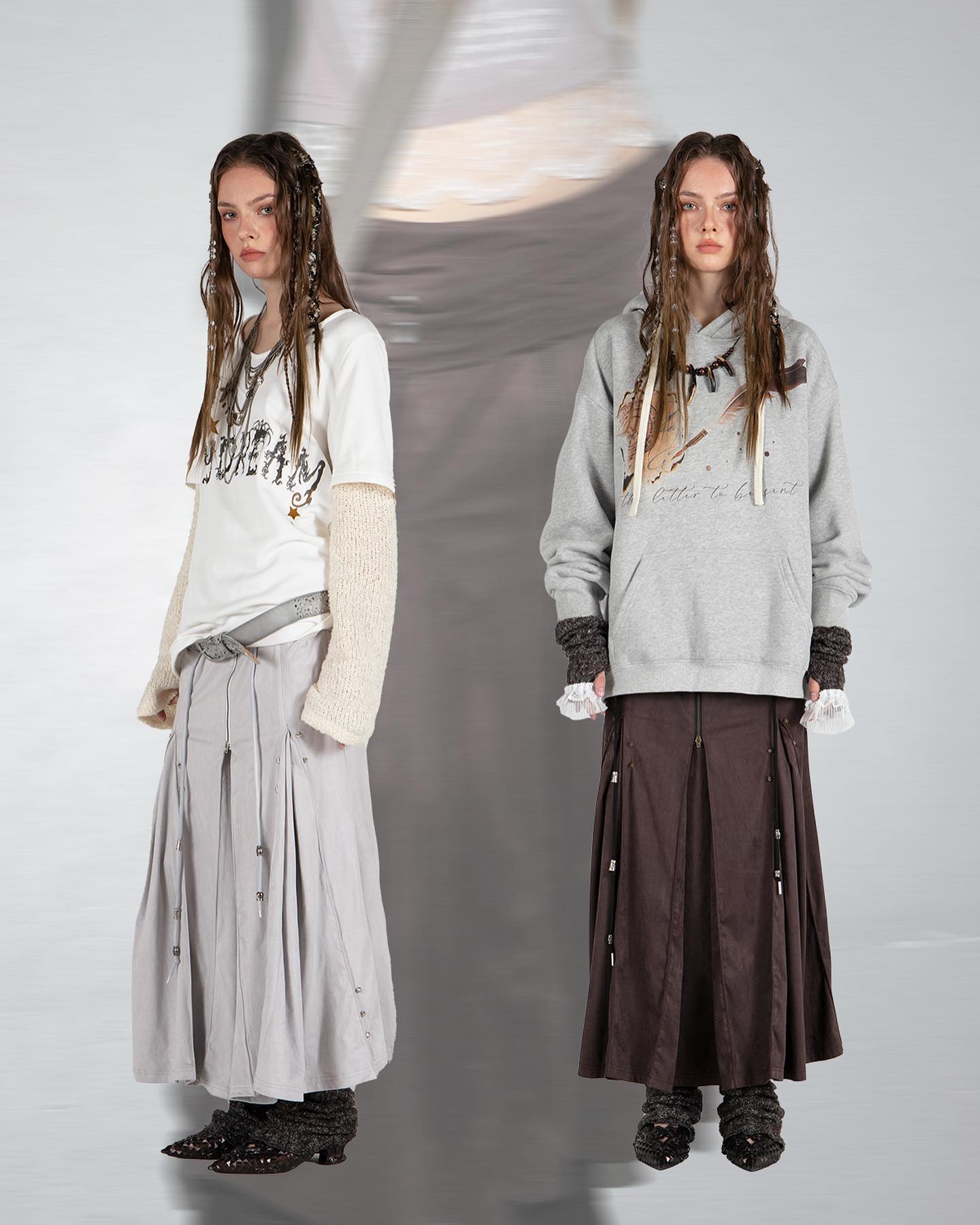 the image showcases two female models dressed in distinct yet coordinated outfits. one model wears a creamcolored oversized longsleeved tshirt with a graphic print and a long, pleated grey skirt. the other model sports a grey hoodie with a graphic design and a brown pleated skirt. both skirts are long and reach the ankles, falling in gentle pleats.  both outfits are accessorized with necklaces and bracelets. their hair is styled with long braids and they are styled in a similar pose against a neutral light grey backdrop. the photo is a fulllength studio shot capturing the details of the clothing. the overall aesthetic is casual yet stylish with a blend of streetwear and more formal elements.  a blurred image appears to be in the background, possibly a moving image from a video or a stylized photographic effect.