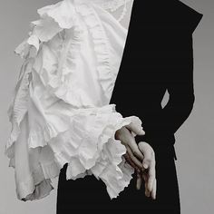 a close up portrait of a model wearing a white blouse with frilled sleeves paired with a black jacket. the sleeves are dramatically frilled, creating a stark contrast against the black jacket. the model's hands are clasped together, their pose further emphasizing the juxtaposition of the black and white. the lighting and overall feel of the image are stark and dramatic, highlighting the model's elegance and the sophistication of the outfit. the highcontrast monochrome color scheme adds a sense of timeless beauty and sophistication.