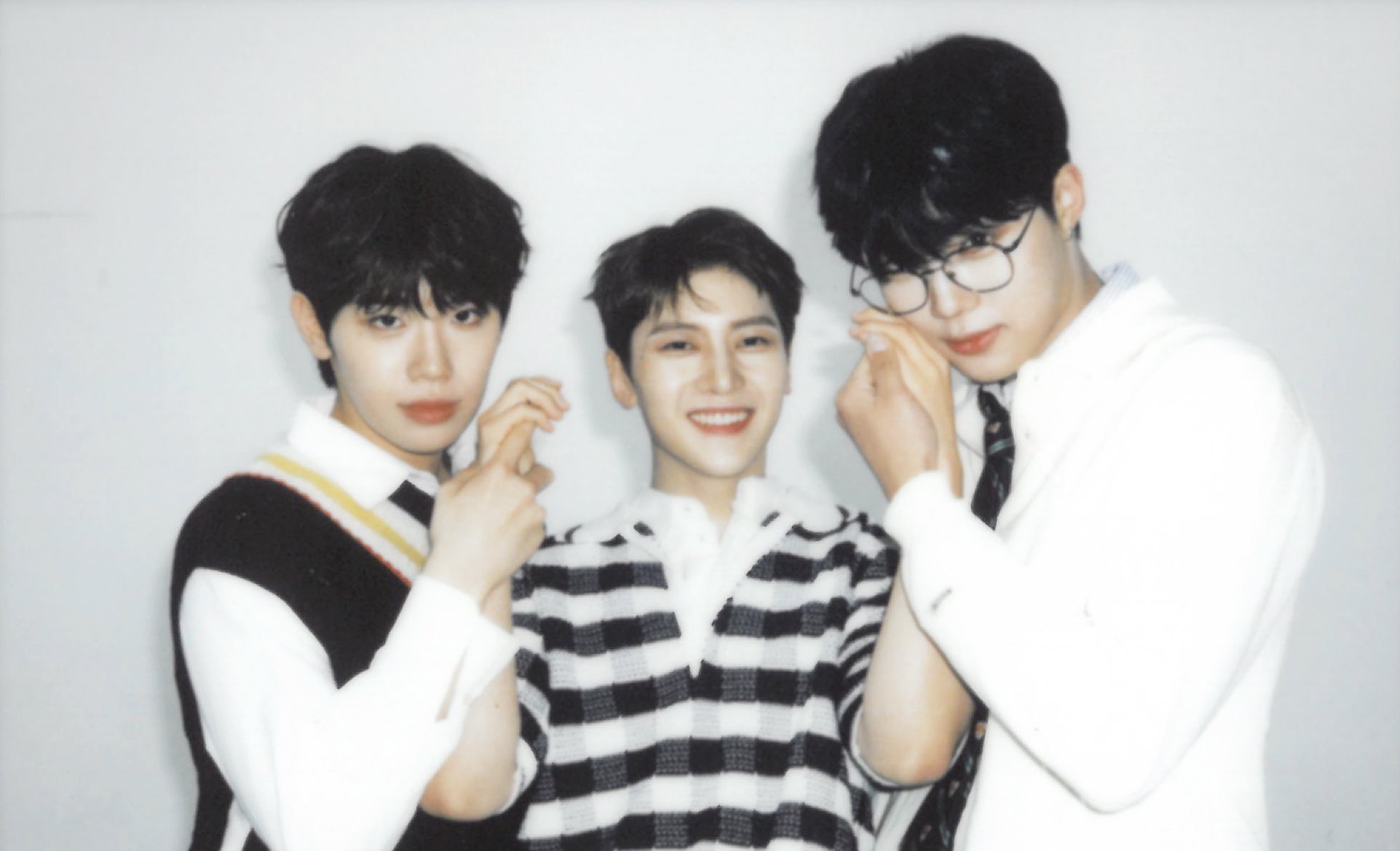 a brightly lit, eyelevel photo of three young men in casual clothing stand close, with their arms intertwined and smiling gently at the camera. the background is plain and white, emphasizing the subjects of the image. the overall tone is warm and friendly.