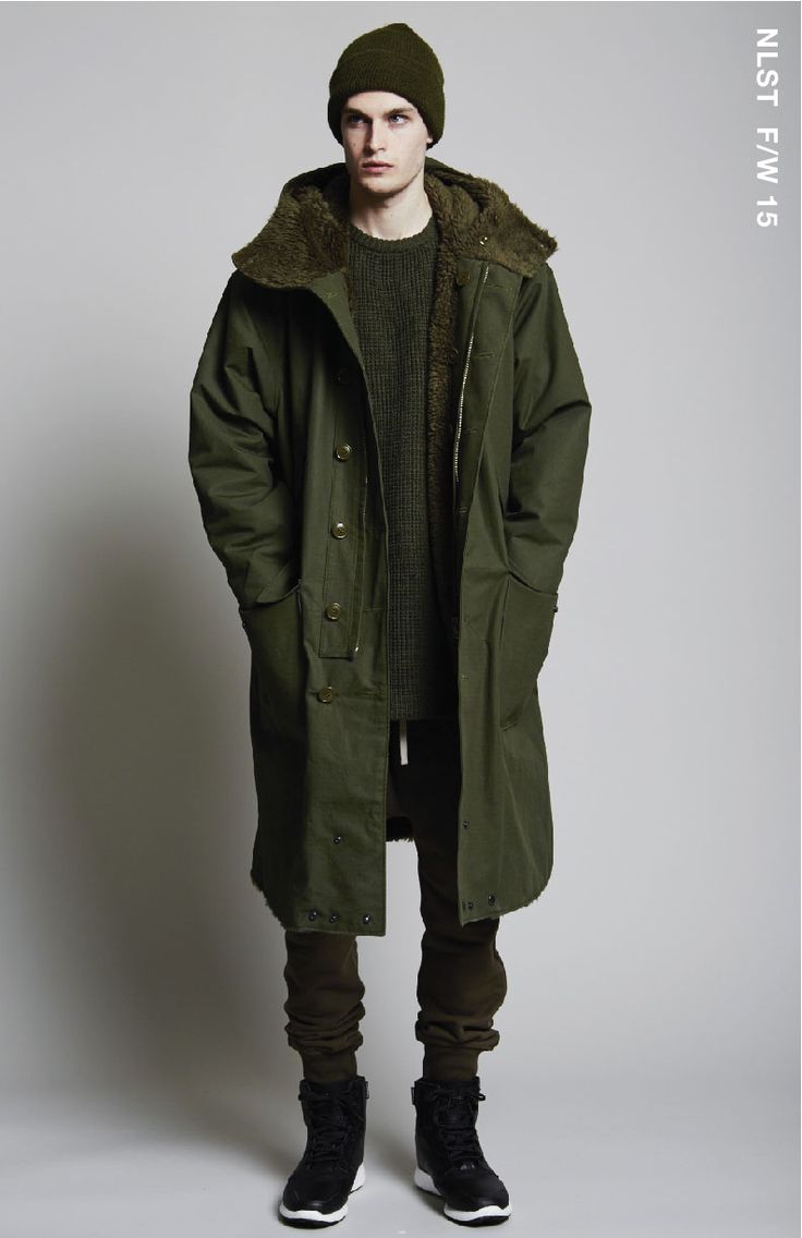 the image showcases a male fashion model wearing an olive green, long parkastyle coat with a sherpalined hood. he's also sporting a matching olive green beanie, dark olive green sweatpants, and black hightop sneakers.  the model stands against a plain light grey background. the image is a professional studio shot, likely from a fashion catalog or lookbook, as indicated by the "nlst f/w 15" label, suggesting a clothing brand's fall/winter 2015 collection. the overall style is casual yet sophisticated, with a hint of militaryinspired design in the coat.
