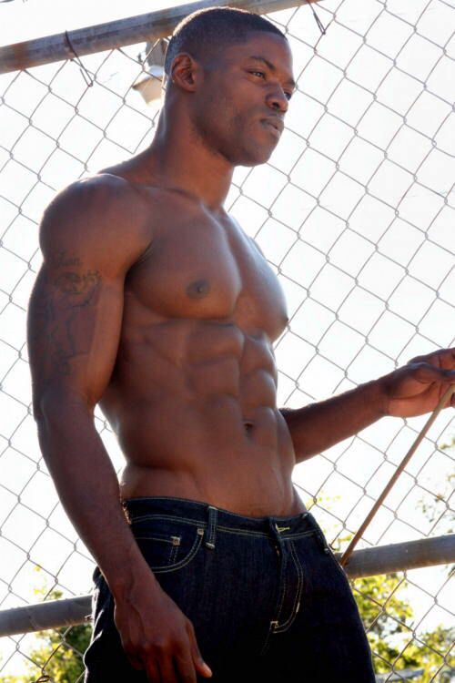 a shirtless muscular man with a six pack is standing outdoors, looking away from the camera with a smile. he is wearing blue jeans and leaning against a chain link fence.  the photo is taken from the side.