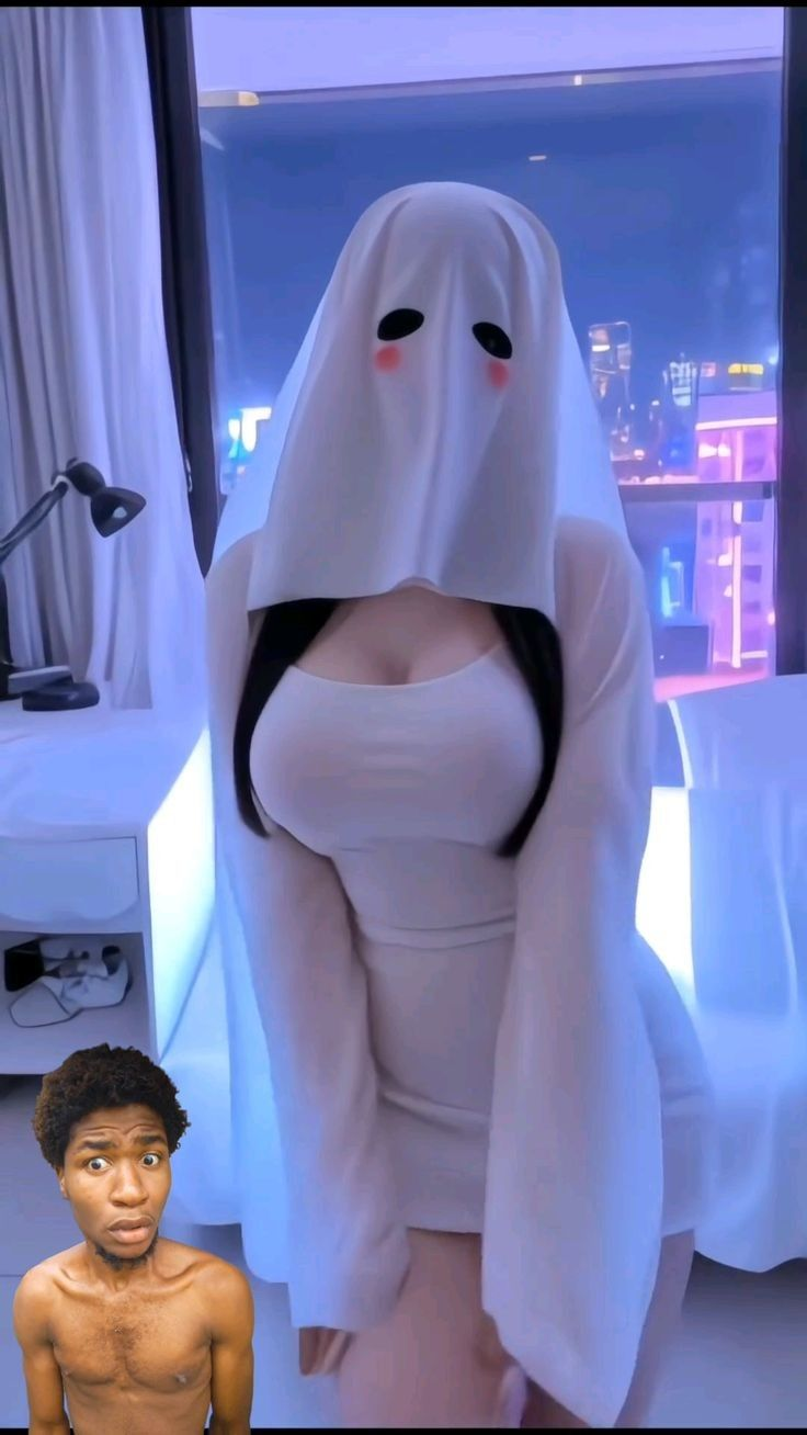 the image shows a person dressed as a ghost with a white sheet and two black eyes, and red cheeks. the person is wearing a white dress and has long black hair. in the bottom left corner is a man with a surprised expression. the background features a window with city lights.