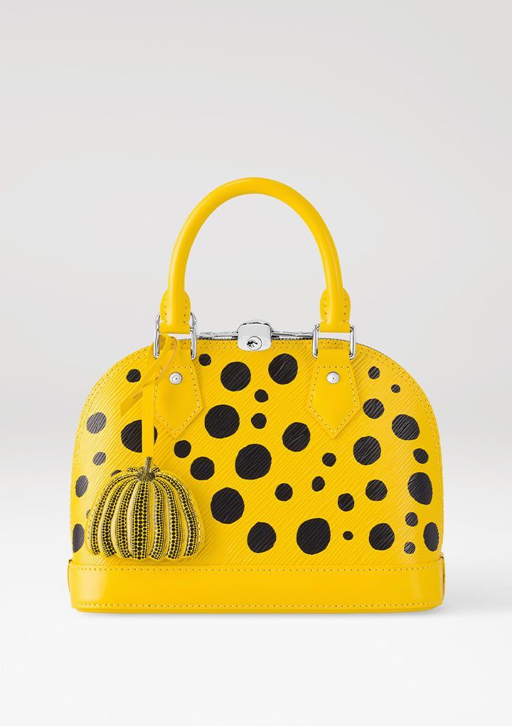a yellow louis vuitton handbag with black polka dots is displayed against a white background. the bag has a rounded shape with a top handle and a small pumpkin charm hanging from the side. the overall style is fashionforward and highend.