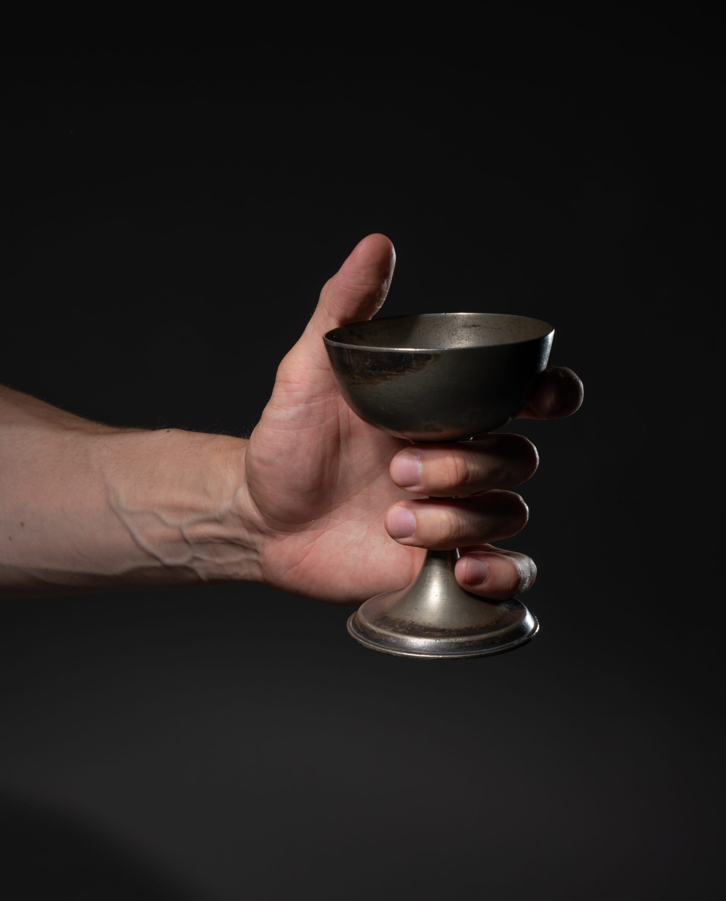 a hand holds a tarnished silver goblet against a black background. the goblet is made of metal and has a rounded bowl and a stem with a circular base. the hand is that of a man and is positioned in the center of the image. the background is black and provides a stark contrast to the goblet. the lighting is soft and highlights the goblet's metallic surface. the overall impression is one of mystery and intrigue.