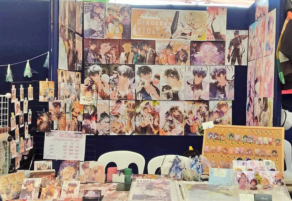 detailed closeup shot captures a vibrant artist's booth at an anime convention. the backdrop features a collage of meticulously arranged animestyle artwork, predominantly showcasing characters with dark hair and expressive eyes. the pieces are diverse in style and composition, adding depth to the overall display.the booth's foreground is equally engaging, boasting an array of merchandise including keychains and prints featuring the same animeinspired characters. a price list is visible on the table, indicating the cost of the displayed products. the setting is lively and dynamic, conveying the atmosphere of a bustling convention. the booth demonstrates the artist's dedication to presenting their work in an appealing and organized manner.