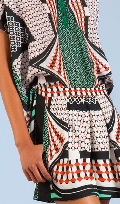 loseup view of a woman's shortsleeved dress featuring a vibrant geometric print in black, white, red, and green. the dress is pleated at the bottom, and the fabric appears to be a smooth silklike material.  the model's arm and hand are partially visible. the background is a muted light blue.