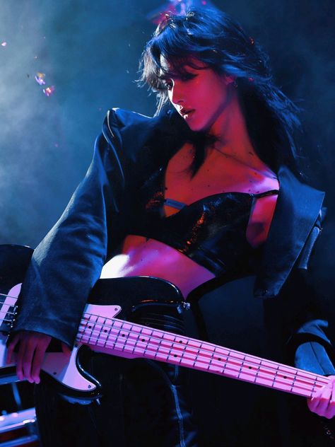 a woman is on stage holding a bass guitar. she is wearing a jacket and a revealing top. stage lights illuminate her and the guitar.