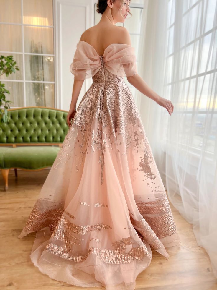 a woman in a beautiful, pink, tulle gown with rose gold embroidery is posing in a studio setting.  the dress is offtheshoulder with a bow at the neckline. the woman is looking off to the side, her back to the camera. the gown has a flowing skirt with a long train. the woman is standing in front of a window with white curtains. there is a green velvet chair to the left of her. the studio floor is made of wood.