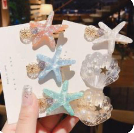 the image showcases a set of six hair clips arranged on two white cards. four of the clips feature a starfish design in pastel pink, blue, and green, adorned with glitter and a faux pearl accent. the remaining two clips are shaped like seashells, crafted from clear acrylic and embellished with a pearl and a small star. the clips are held by a hand with a nude manicure, suggesting they are intended for personal use. the overall aesthetic exudes a feminine, summery, and whimsical charm.