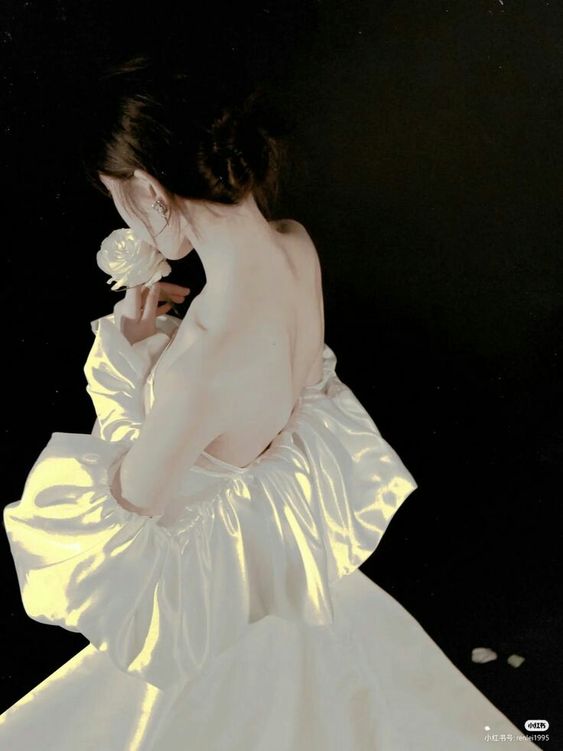 a woman with her back turned towards the camera stands holding a white rose to her face. she is wearing a white offtheshoulder dress with voluminous, ruffled sleeves. the background is dark and creates a dramatic contrast with the woman's pale skin and the white dress. the lighting is soft and diffused, creating a romantic and ethereal atmosphere.