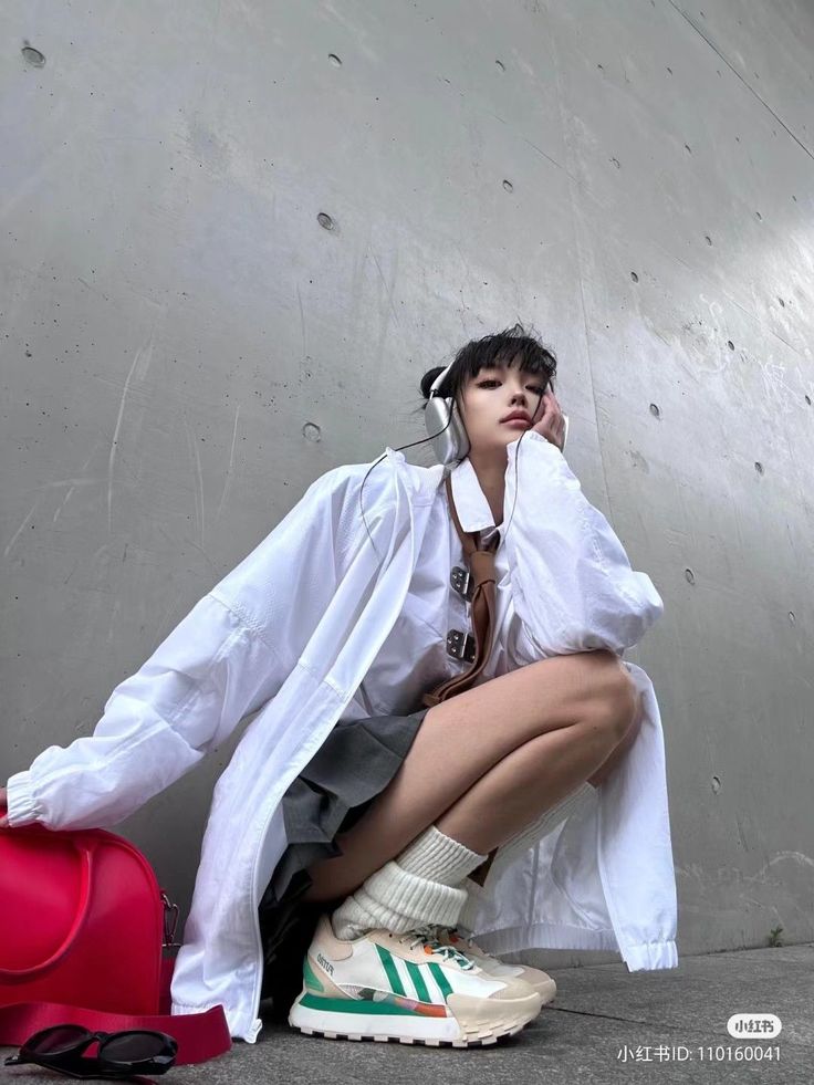 a woman is squatting outdoors, wearing headphones, a white jacket, a gray skirt, and sneakers. she is also carrying a red bag and has sunglasses nearby.