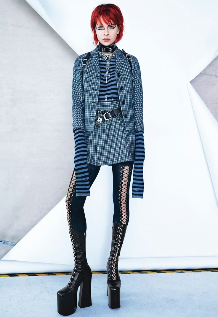 a full shot of a woman with red hair, wearing dark makeup and fashion, standing in front of a white background. she has a punk, goth look, including a striped long sleeve shirt, a plaid jacket and skirt, lacy tights and black platform boots. she is also wearing several pieces of jewelry, including a cross necklace and a choker.