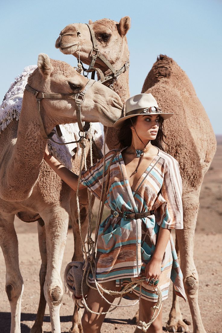 a fashion photograph featuring a woman in a stylish westernboho outfit, standing between two camels in a desert setting. she is wearing a striped turquoise and beige poncho, a brown cowboy hat, a belt with a large buckle, and various jewelry. the camels are harnessed and appear calm. the overall mood is glamorous and adventurous. the setting is a sandy desert under a clear blue sky. the image is professionally shot with good lighting and composition, suggesting a highfashion editorial shoot.
