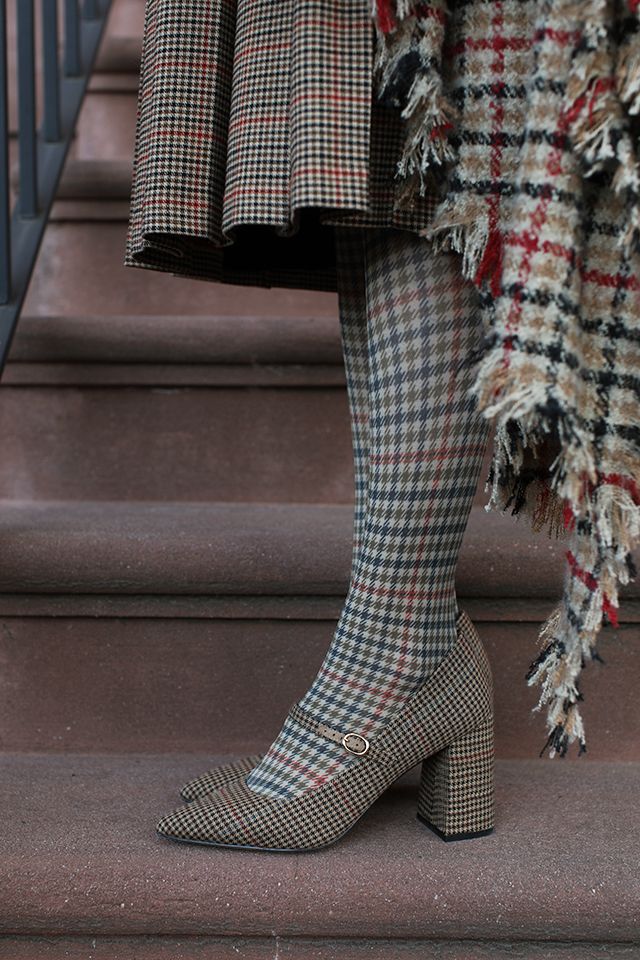 loseup view of a woman's legs wearing plaid tights and plaid shoes. the shoes are mary jane style with a chunky heel. the woman is standing on a brown stone staircase. a plaid fringed scarf is draped around her waist. the overall style is autumnal and sophisticated.