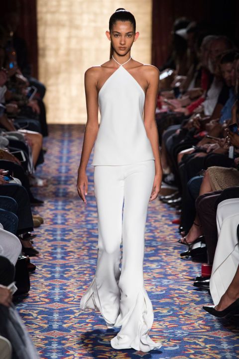 a beautiful model in a white halter top and pants walks the runway. she has on a ponytail and is wearing white high heels. the catwalk is decorated with a colorful patterned rug. there are people seated on either side of the runway.