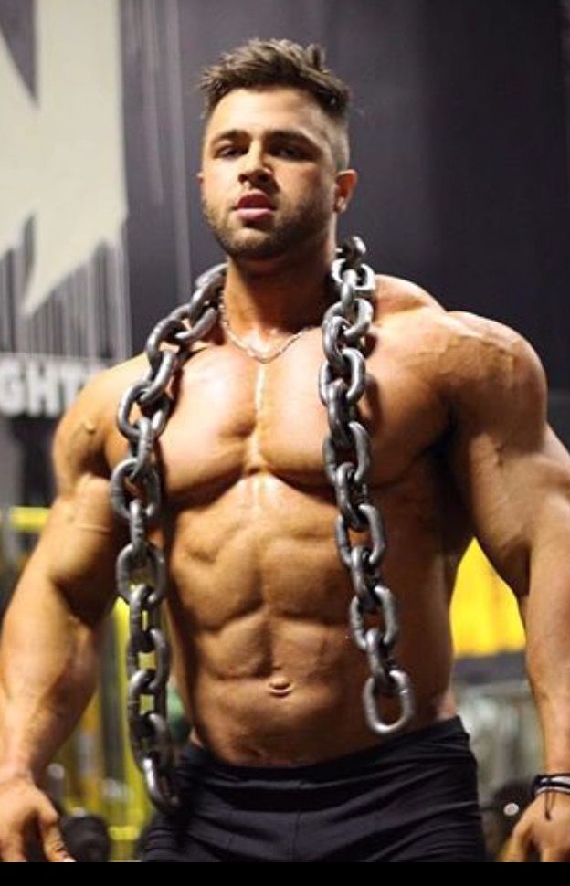 a muscular man with a sixpack, wearing black shorts, poses with a chain wrapped around his neck and shoulders. he is in a gym setting, with a dark background, and he is looking at the camera with a serious expression. he appears to be in very good shape.