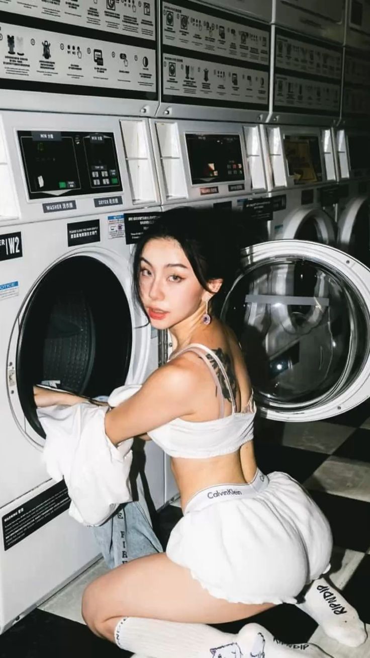 a woman is kneeling in a laundromat in front of a row of washing machines. she's wearing a white cropped top and white calvin klein shorts with white ripndip socks. her hair is up, and she has an earring and a tattoo on her back. she is loading clothes into a washing machine.