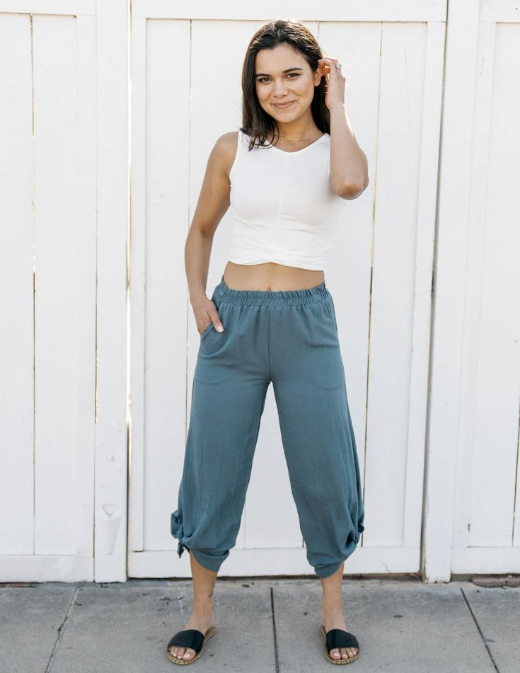 a young woman is standing in front of a white wall, posing for the camera. she is wearing a white tank top, blue pants, and brown sandals. her outfit is casual and stylish, perfect for a summer day. the background is a simple white wall, which helps to focus the attention on the woman and her outfit.