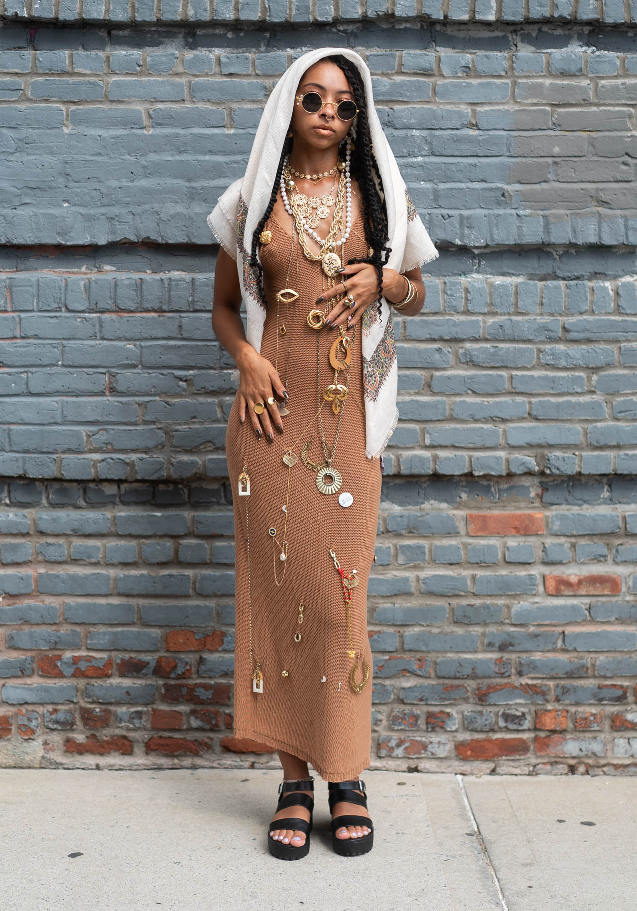 a woman wearing a brown dress, a white shawl, and many gold necklaces poses in front of a brick wall. she is wearing sunglasses and black sandals.