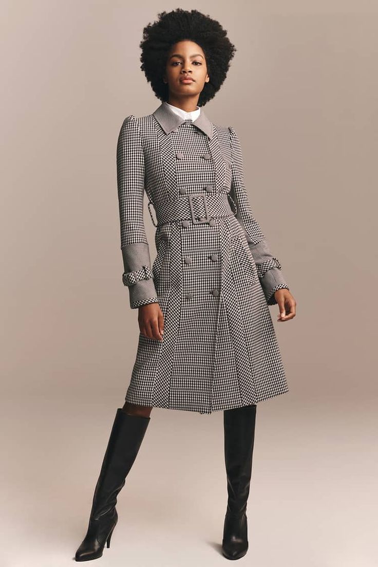 a woman is posing in a studio wearing a black and white gingham trench coat with grey accents and black knee high boots. she is standing with a slight pose and her arms are slightly out to the side.