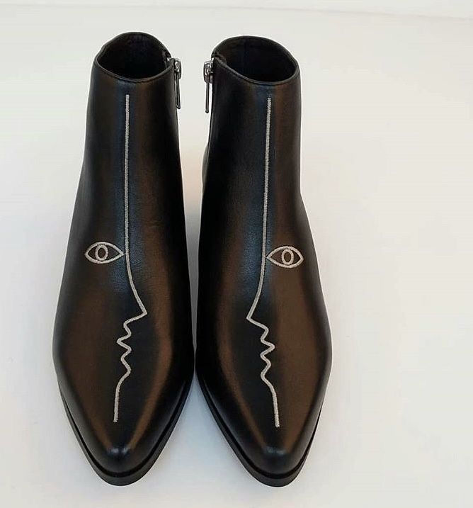 a pair of black leather ankle boots with pointed toes are shown in the image.  the boots feature a minimalist design with a subtle line drawing of a face, including an eye, embroidered on each boot in a lightcolored thread.  the boots are positioned on a white background.