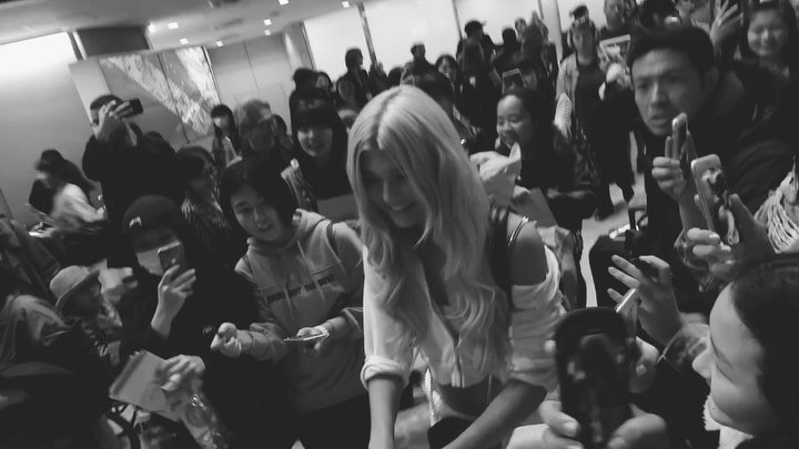 a black and white image shows a crowd of people, many holding up smartphones to take pictures or videos. a celebrity with blonde hair is signing autographs for fans. the scene suggests an event or public appearance where the celebrity is interacting with their fans.