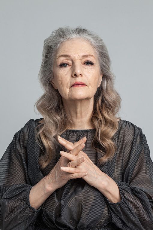 a portrait of an older woman with gray hair. she has her hands clasped in front of her. she is wearing a black blouse and has a serious expression.