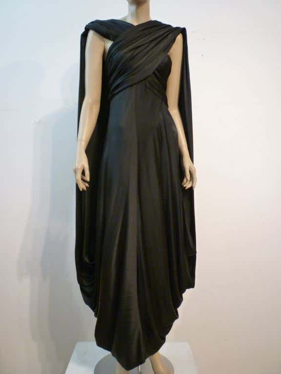 a fulllength studio shot of a black evening gown on a mannequin. the dress features a draped cowl neckline and a capelike overlay that adds volume and movement. the fabric appears to be a soft jersey or knit material. the dress is fulllength and flows to the floor, creating a dramatic silhouette. the overall style is sophisticated and timeless.
