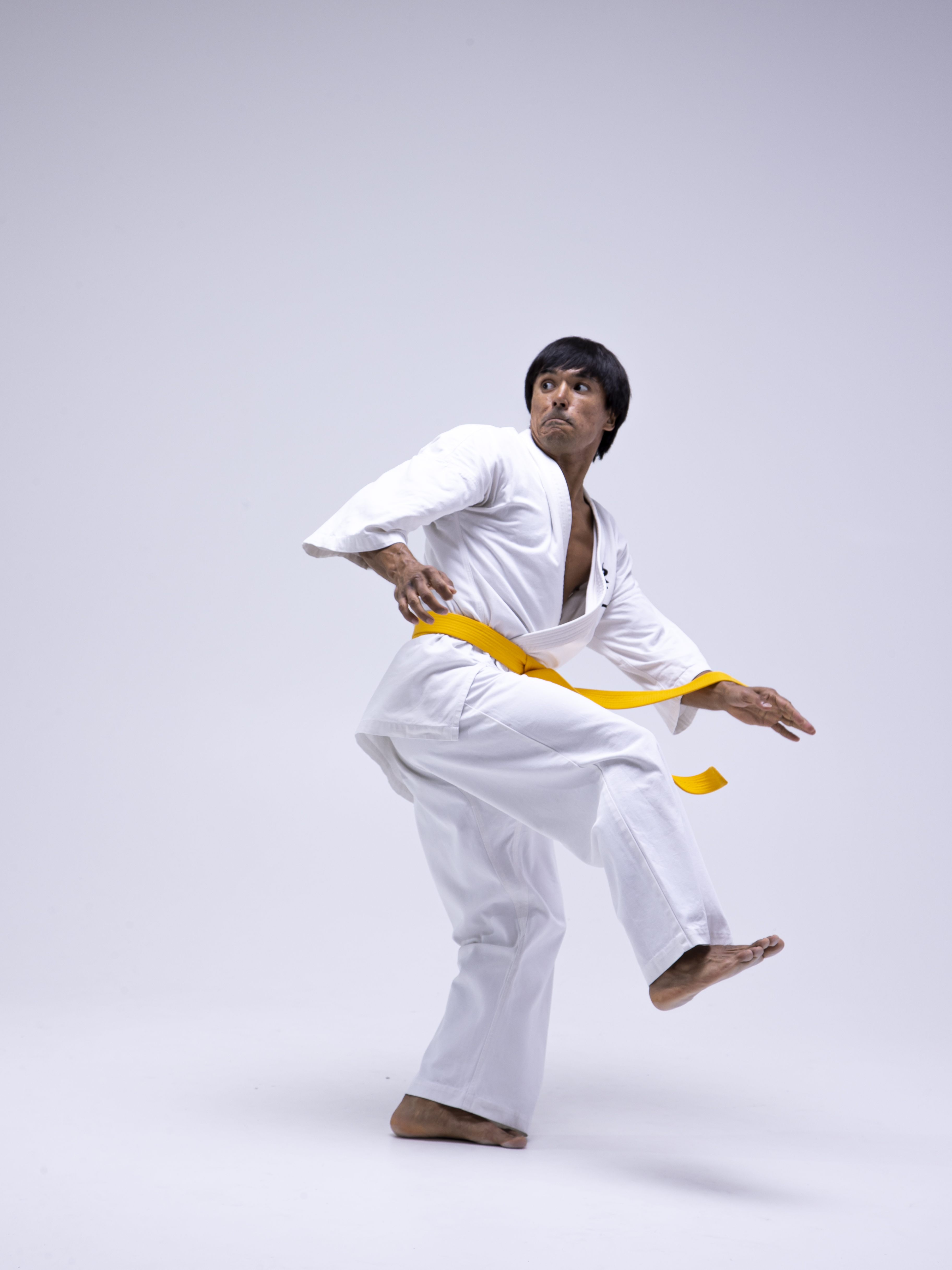 a man in a white karate uniform with a yellow belt is performing a kick in what appears to be a dojo. the background is plain white. he is barefoot and looking to his left.