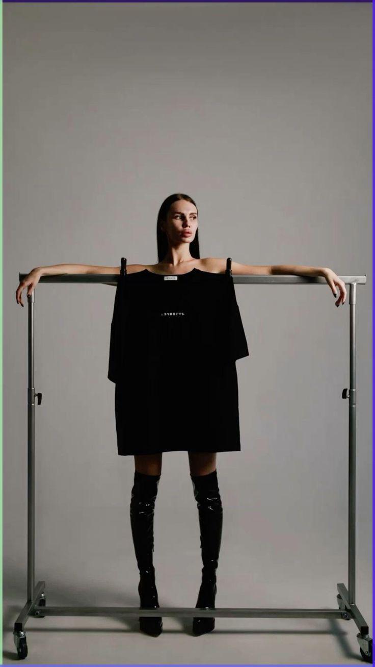 a full shot of a slender woman with long dark hair modeling an oversized black tshirt dress and black thighhigh boots. she is standing on a simple metal clothing rack against a plain gray background. the overall aesthetic is minimalist and elegant.
