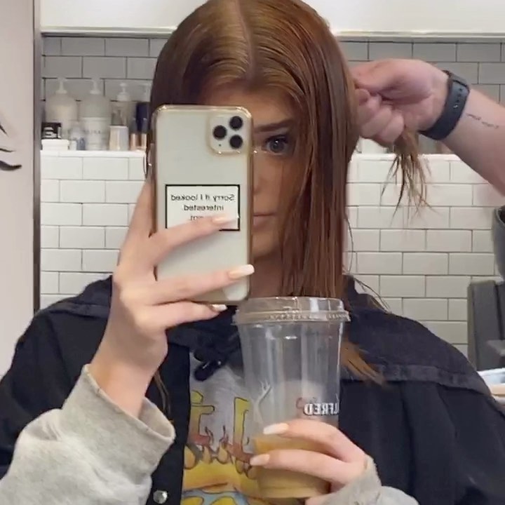 a woman holding her cellphone and a drink in what appears to be a hair salon or barbershop. a hairstylist is cutting a strand of her hair. she is wearing a black top and a grey sweatshirt. the wall behind her is made of white tiles.
