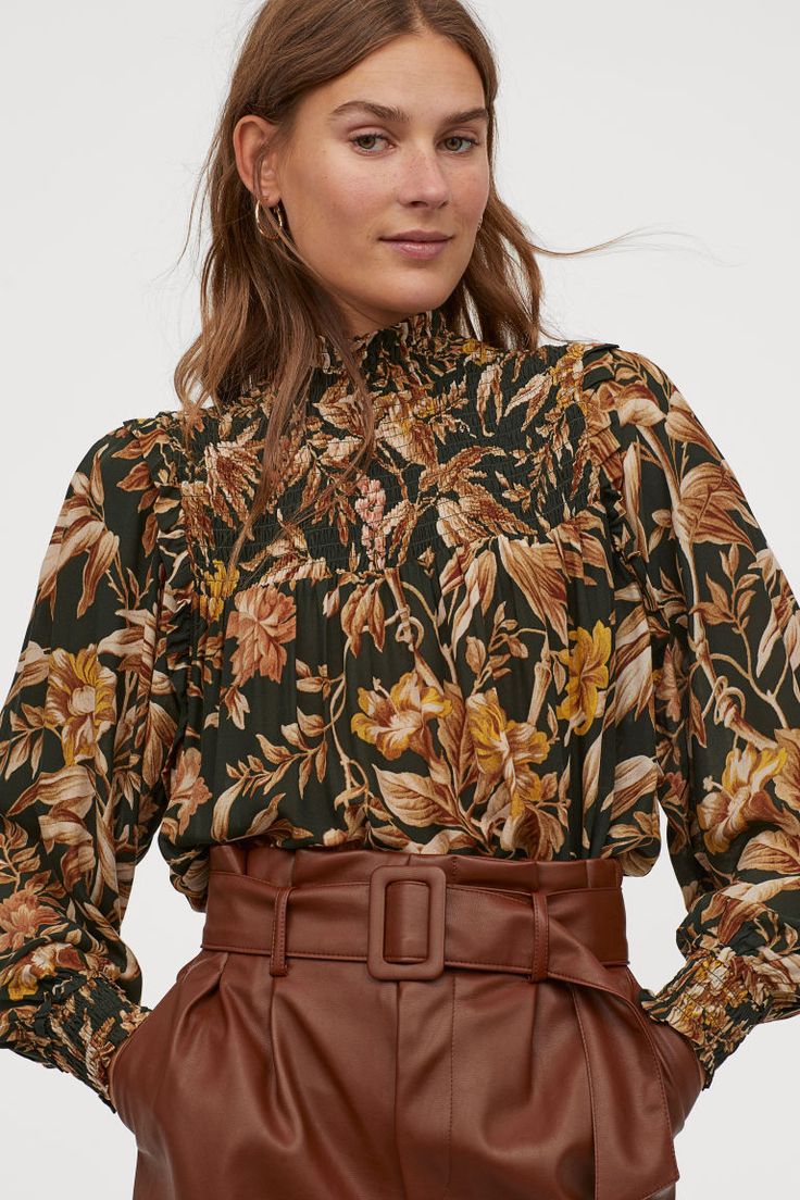 closeup view of a woman wearing a dark green floral blouse with a high neck and smocked detailing. the blouse has long sleeves and a fitted silhouette. she is also wearing brown leather highwaisted pants with a belt. the woman has long brown hair and is looking slightly towards the camera. the image is welllit and the background is a simple, lightcolored backdrop. the style is autumnal or fall fashion.