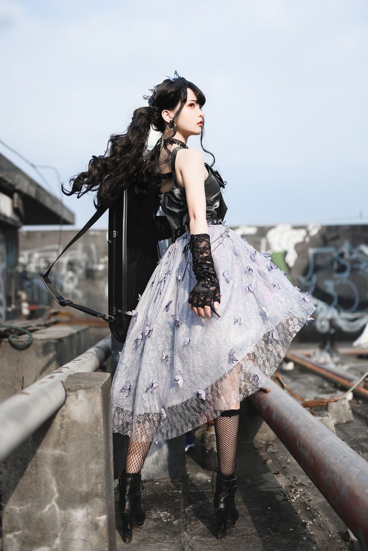 a young woman stands outdoors on a rooftop in a striking cosplay outfit. she is posed in a threequarter view, looking off to the side with a contemplative expression. her long, black hair is styled in a high ponytail, and she wears a small crown or decorative hairpiece.her outfit consists of a sleeveless black top, paired with an asymmetrical silver and blue skirt that appears to be adorned with butterfly details. she has on black lace gloves and black fishnet stockings. her footwear consists of black platform boots with laceup detailing.the background shows an urban rooftop setting, with graffiticovered walls and various architectural elements under a somewhat cloudy sky. the lighting is soft, suggesting it might be a partly cloudy day.