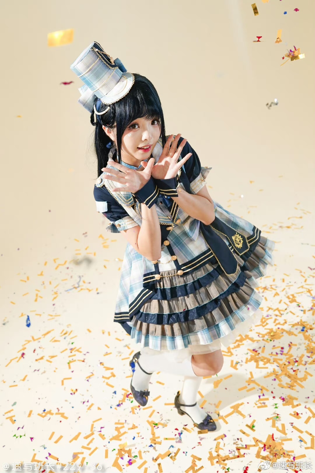young woman is cosplaying an anime idol character in a studio setting. she is wearing a blue and white checkered dress with gold accents and a matching hat. the dress is layered with ruffles and has a large bow on the back. she is also wearing white kneehigh socks and blue shoes. her hair is black and styled in pigtails. she is posing with her hands raised and a cheerful expression on her face. the background is a light yellow color and there is confetti scattered on the floor. the lighting is bright and artificial, giving the image a cheerful and vibrant feel.