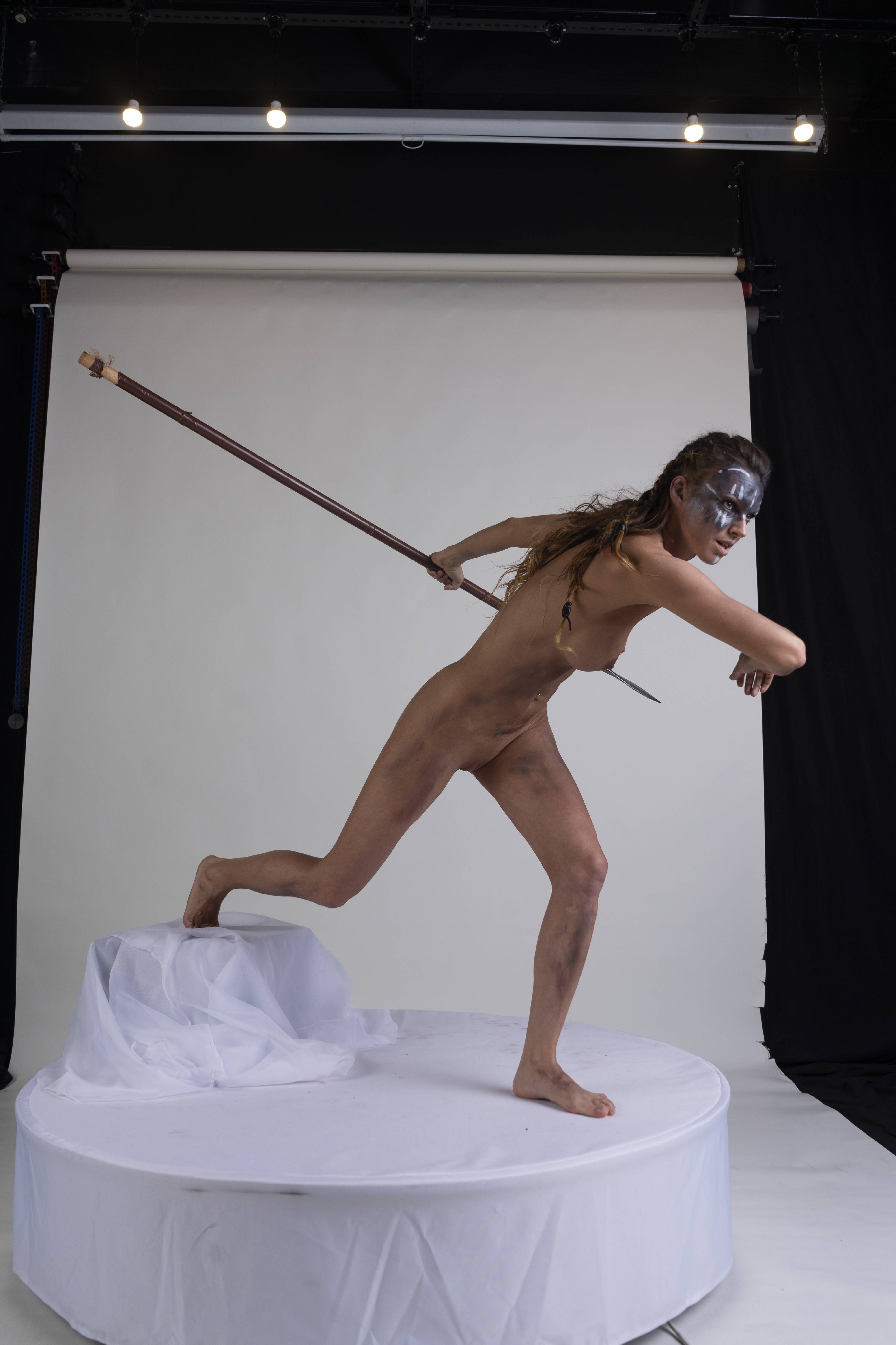 a nude woman stands on a round platform, holding a spear as if throwing it. she has silver face paint on her face and her skin is smudged with dirt. she is running and looks focused. the platform is covered in white cloth, and there is studio lighting in the background.