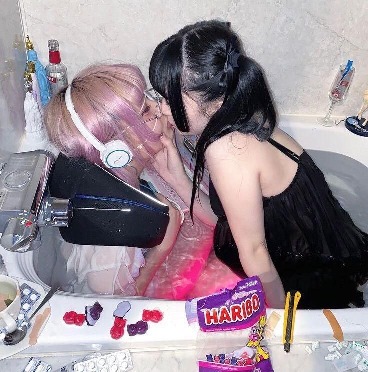 the image depicts two young women in a bathtub. one has pink hair and is wearing headphones, while the other has black hair and a black dress. they are kissing, and the bathtub is filled with pinktinged water.  around the tub are various items including haribo candy, pills, a razor blade, and an electronic device, creating a scene that combines elements of pastel goth aesthetics and potentially suggestive themes. the overall mood is intimate and slightly unsettling due to the juxtaposition of romantic imagery with seemingly unrelated objects.