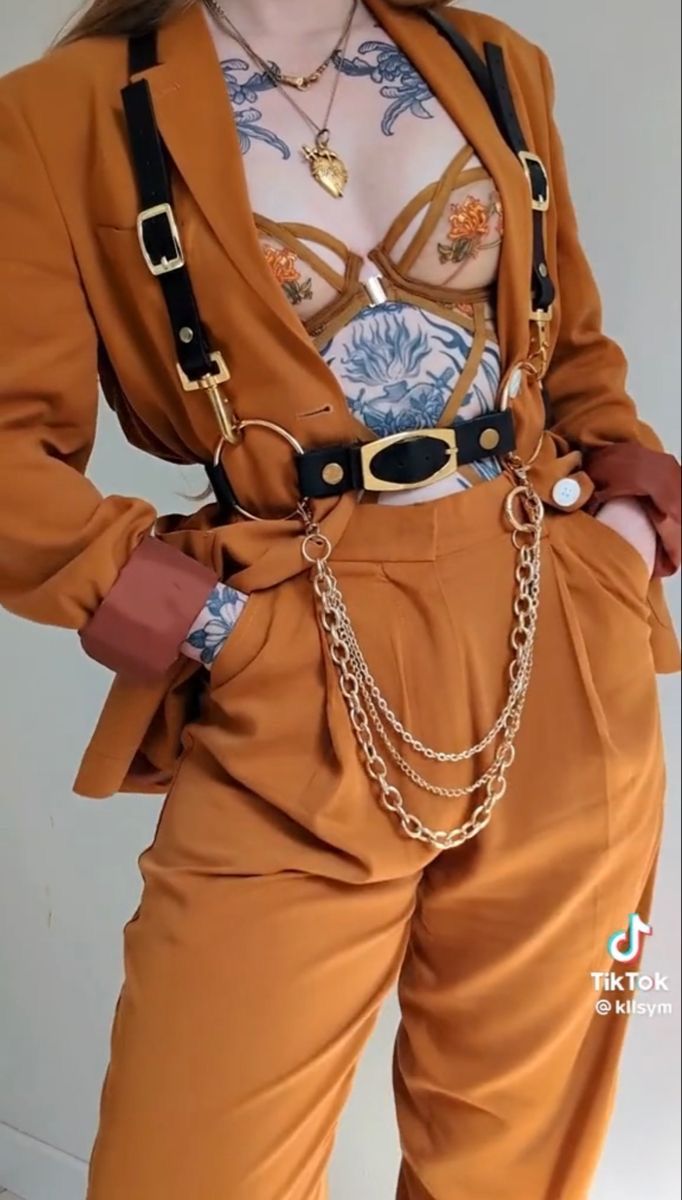 a woman wearing a brown blazer and matching pants, with a gold chain belt and suspenders. she has tattoos on her chest and arms, and is wearing a nude bra with floral embroidery. her hair is parted in the middle and falls to her shoulders. she is looking away from the camera. the image is welllit and the colors are vibrant.
