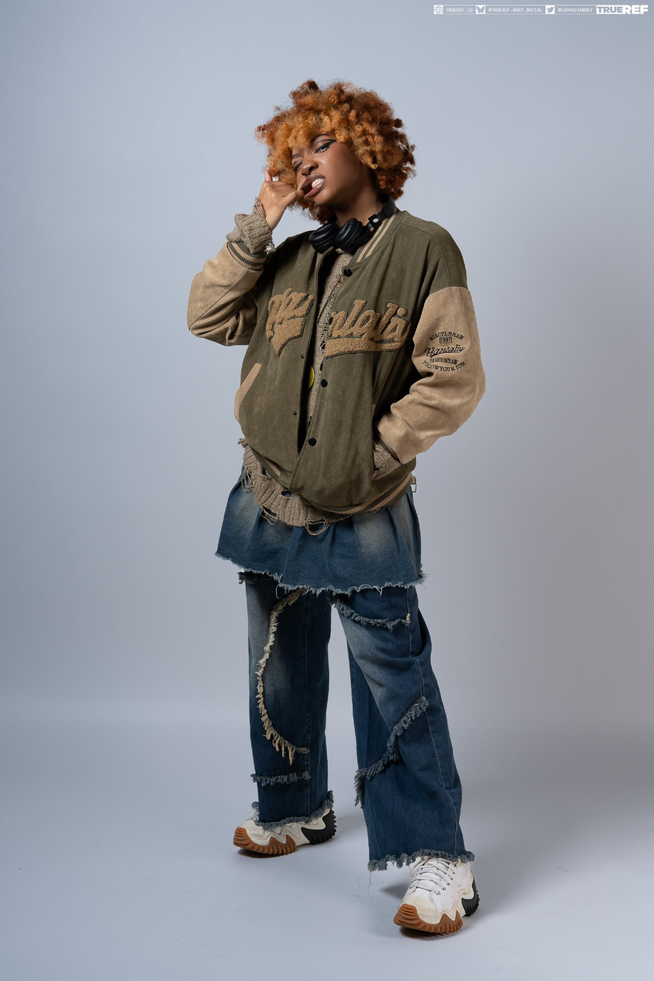 a fullbody studio shot of a young woman with vibrant orange curly hair. she's wearing a stylish outfit featuring a beige and olive green varsity jacket, distressed denim jeans with frayed edges, and white sneakers.  her look is complemented by headphones around her neck. she is posing with a hand near her mouth, displaying a fashionable and somewhat edgy style. the backdrop is a simple light gray, ensuring the focus remains on the model and her attire.