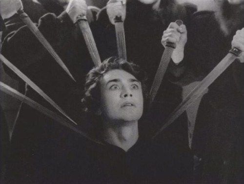 the black and white photograph depicts a man with dark hair, his face conveying a mixture of fear and shock. he's the central figure, seemingly trapped amidst numerous individuals surrounding him, each holding a knife or dagger pointed towards the man. the blades converge on him creating a striking visual effect.  the overall mood is intense, dark, and suggestive of violence or impending doom. the image’s style and composition recall elements of surrealism and cinematic suspense. the high contrast and careful lighting enhance the dramatic effect, making it a powerful and unsettling visual.