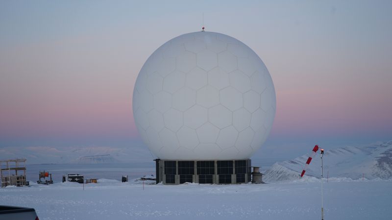 a large white radar dome sits on a snowy landscape. the sky is a light purple and pink. there are mountains in the distance. the dome is part of a weather station or a radar station.