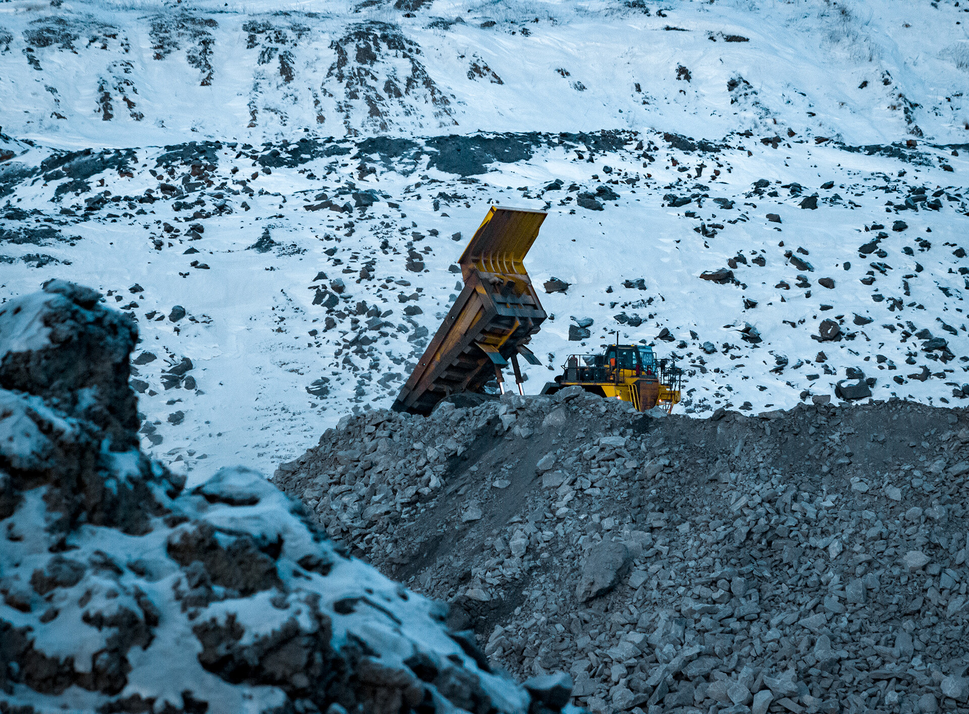 a yellow dump truck is partially buried in a pile of black gravel. the truck is tipped up,  with its bed raised and its tires visible. there is snow on the ground and on the rocks around the truck, which suggests that the location is a quarry or mine site. the truck is likely being used to transport material to another location. the truck is likely a part of a larger operation, and the truck is likely being used in conjunction with other heavy equipment. the image is taken during the day. the sky is overcast and the light is diffused. the photo is taken from a low angle, and it is slightly zoomed in. the image is wellcomposed. the colors are vivid and the details are sharp.