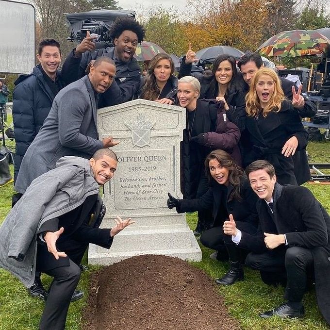 the image shows the cast of arrow gathered around oliver queen's tombstone. the characters are in black and dark clothing and appear somber and grieving. the tombstone reads "oliver queen 19852019" with the inscription "beloved son, brother, husband, and father. hero of star city. the green arrow." the characters include felicity smoak, laurel lance, john diggle, and the flash.